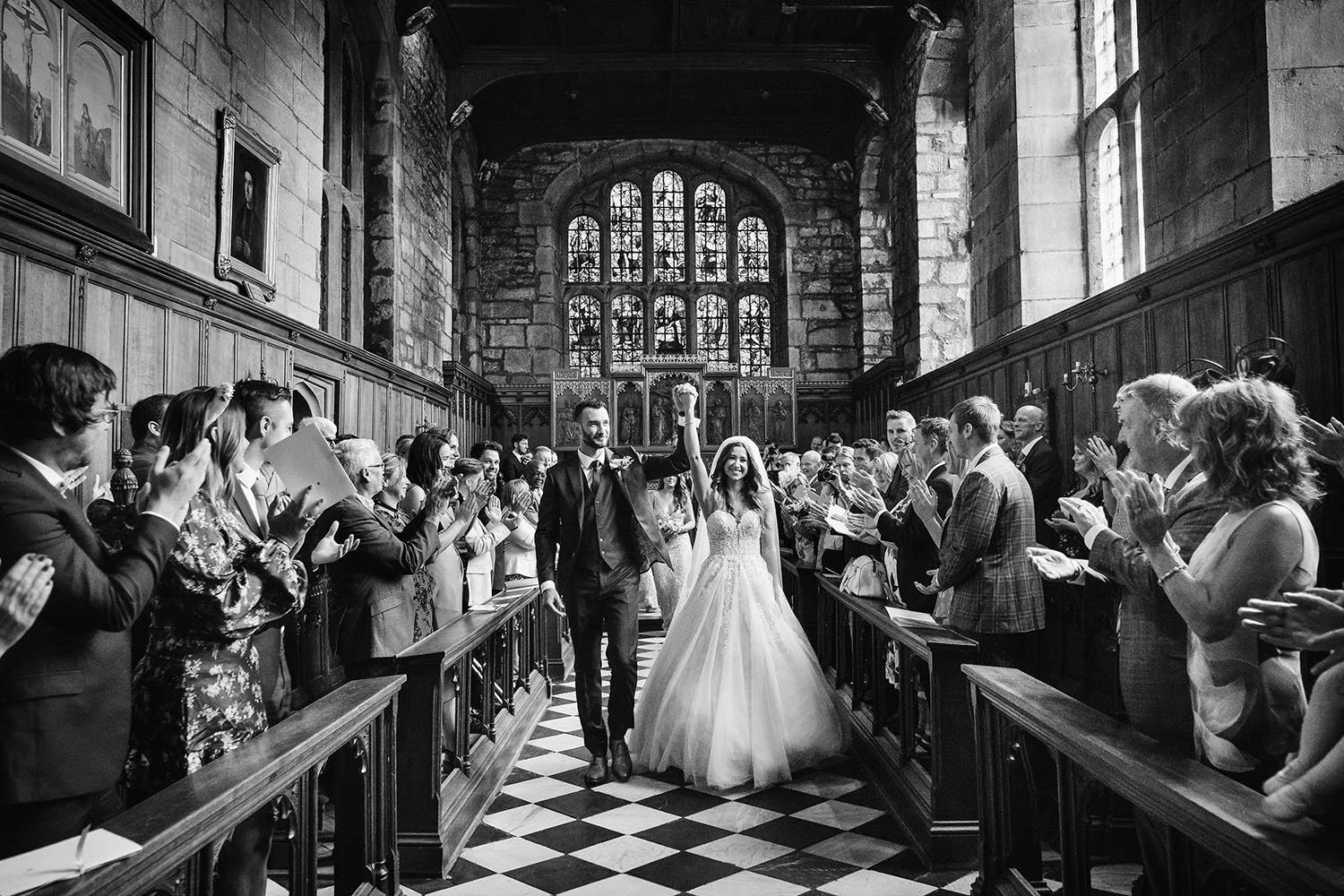 Durham Castle wedding: bride and groom walking back down the aisle of a college chapel as guests applaud, black and white