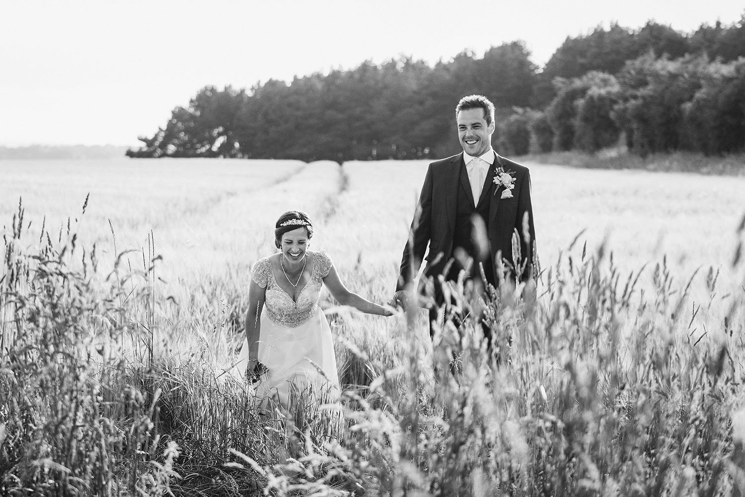Healey Barn wedding: bride and groom walking hand in hand through a wheat field, in black and white.
