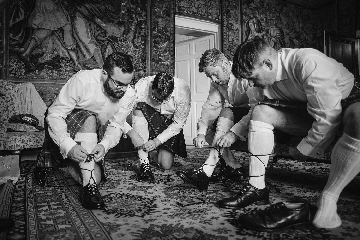 Durham Castle wedding: groom and groomsmen in kilts lacing their shoes while getting ready, in black and white.