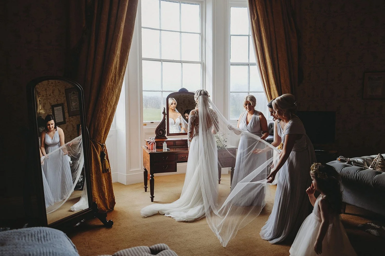 Bride in a wedding gown and long veil prepares with bridesmaids in a sunlit bedroom.