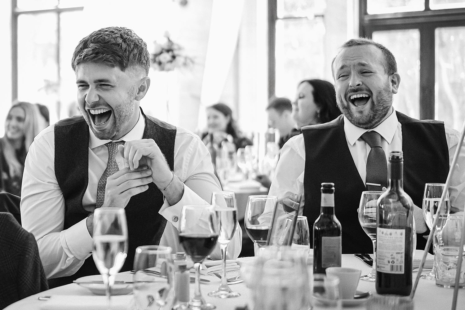 Beamish Hall wedding: two men in waistcoats laughing out loud at the wedding breakfast among wine glasses, in black and white.