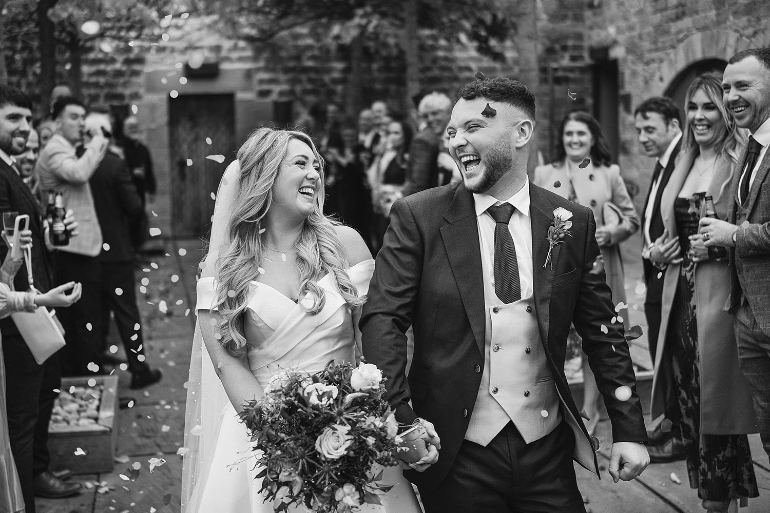 Black-and-white wedding photo of a laughing bride and groom walking through a confetti-filled crowd outside a stone building.