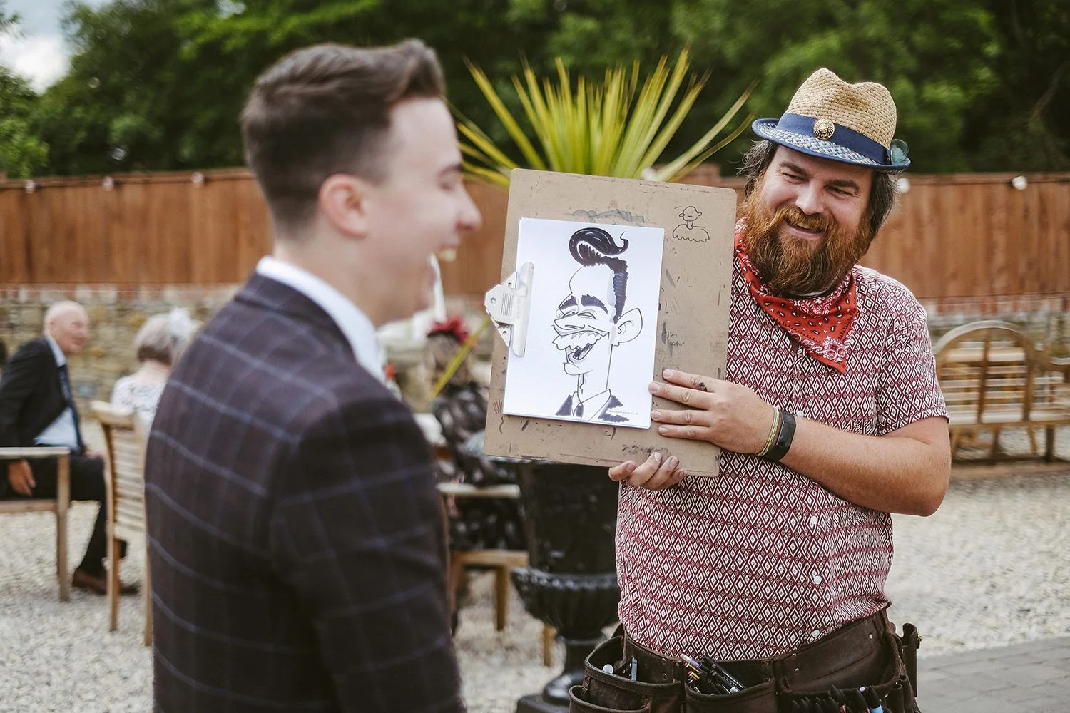 South Causey Inn wedding: groom laughing as a caricature artist in a straw hat shows off a cartoon drawing of him.