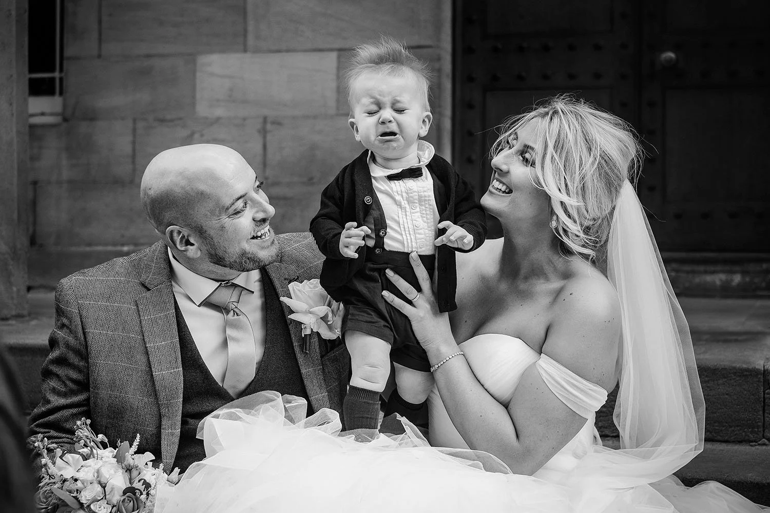 The Vermont Hotel wedding: smiling bride and groom with a crying baby boy in a tuxedo between them, black and white