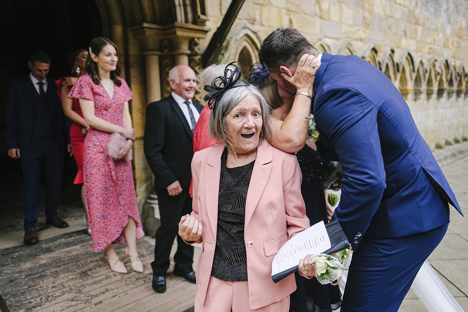 Brinkburn Priory wedding: elderly guest in a pink suit looking delighted as the groom kisses her, outside a stone church