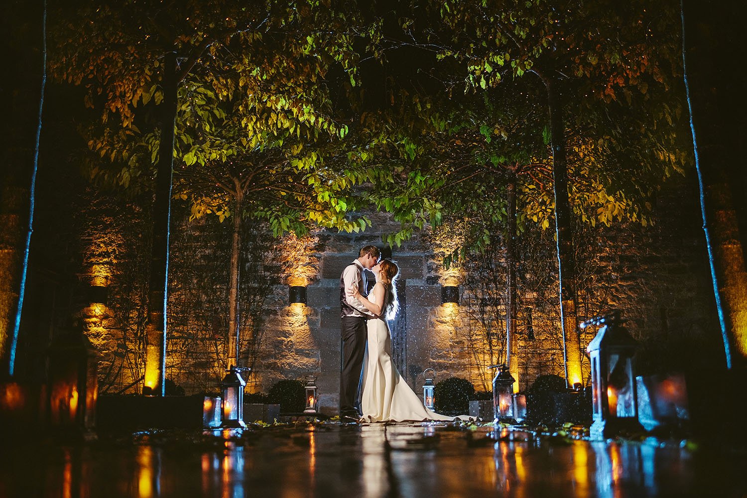 Healey Barn wedding: bride and groom kissing in a stone courtyard lit by lanterns and uplighters among tall trees at night