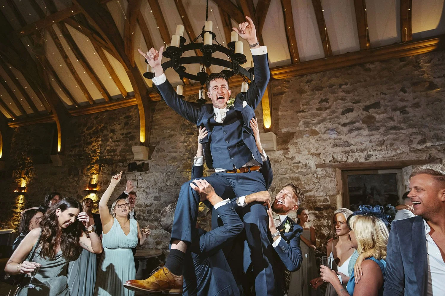 Healey Barn wedding: groom lifted into the air by groomsmen as guests cheer in a stone barn.