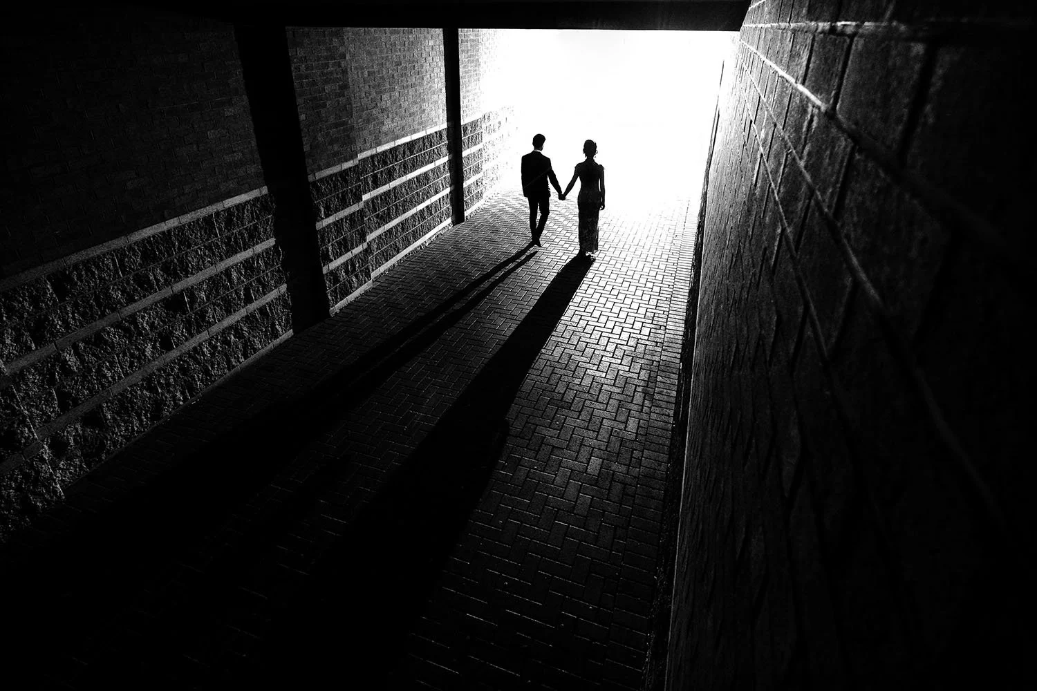 True Jesus Church wedding: bride and groom walking hand in hand as silhouettes through a brick tunnel, black and white