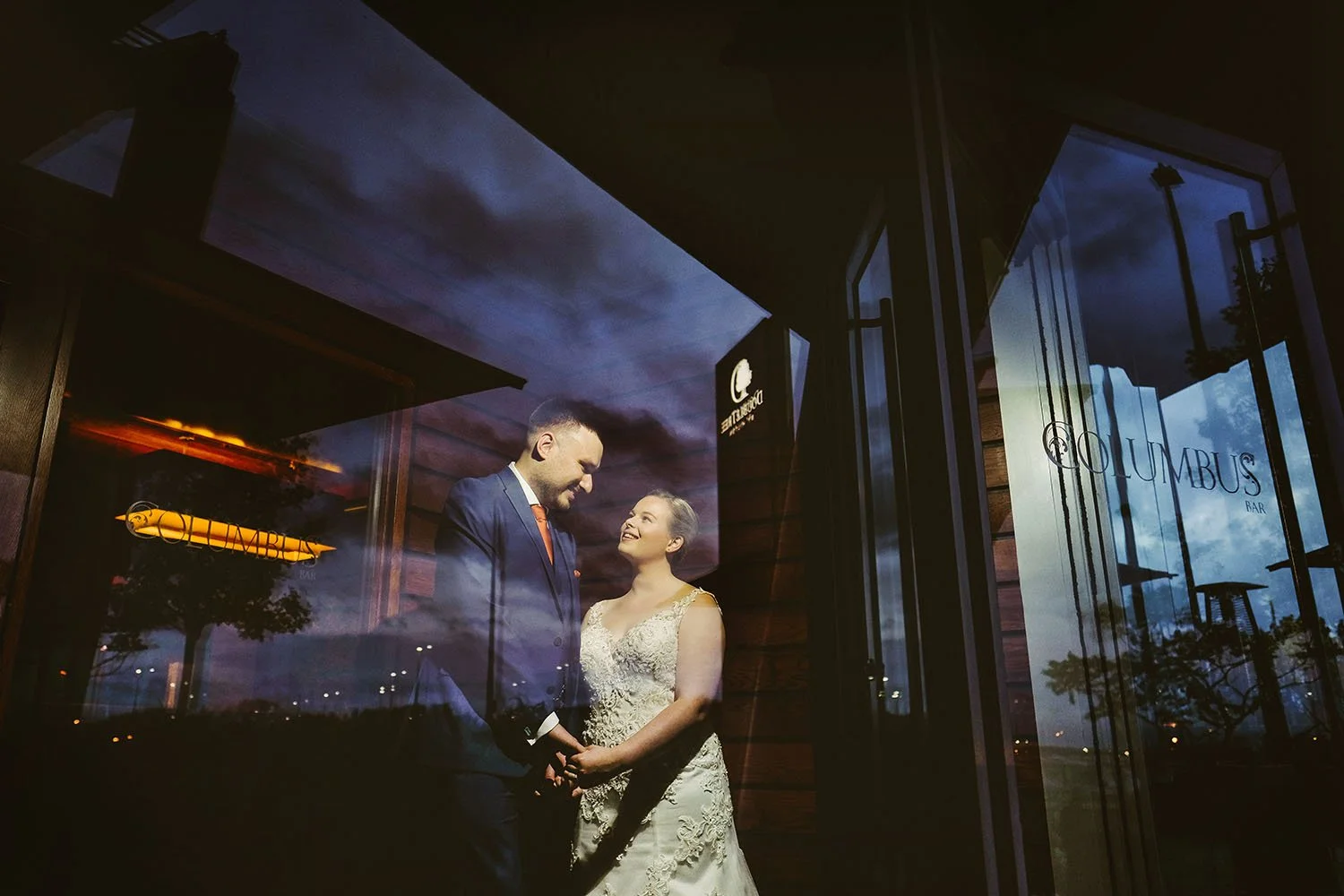 Newcastle wedding: bride and groom holding hands in front of large glass doors reflecting a dusk sky