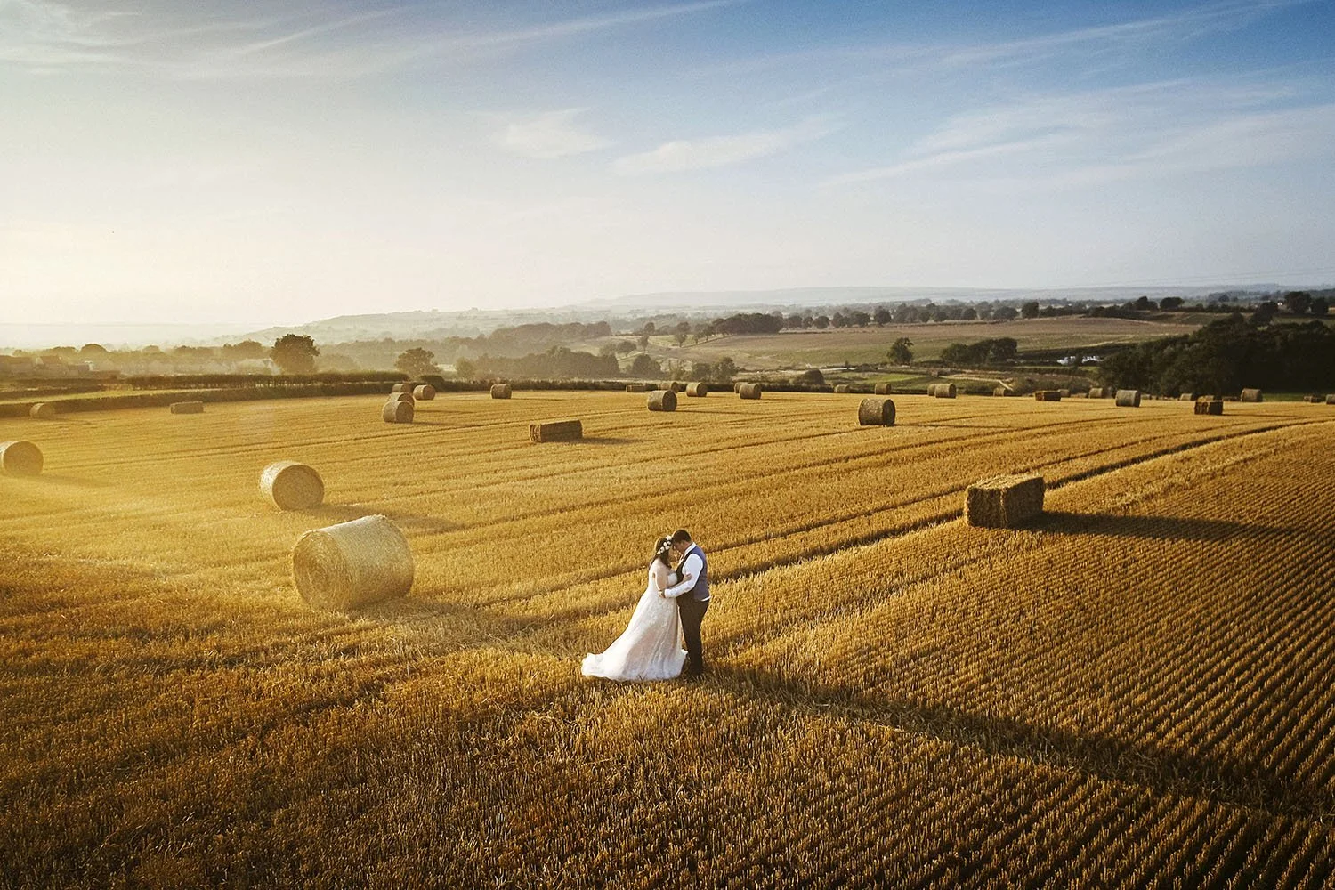 Newlywed couple embracing in a sunlit harvested wheat field surrounded by hay bales