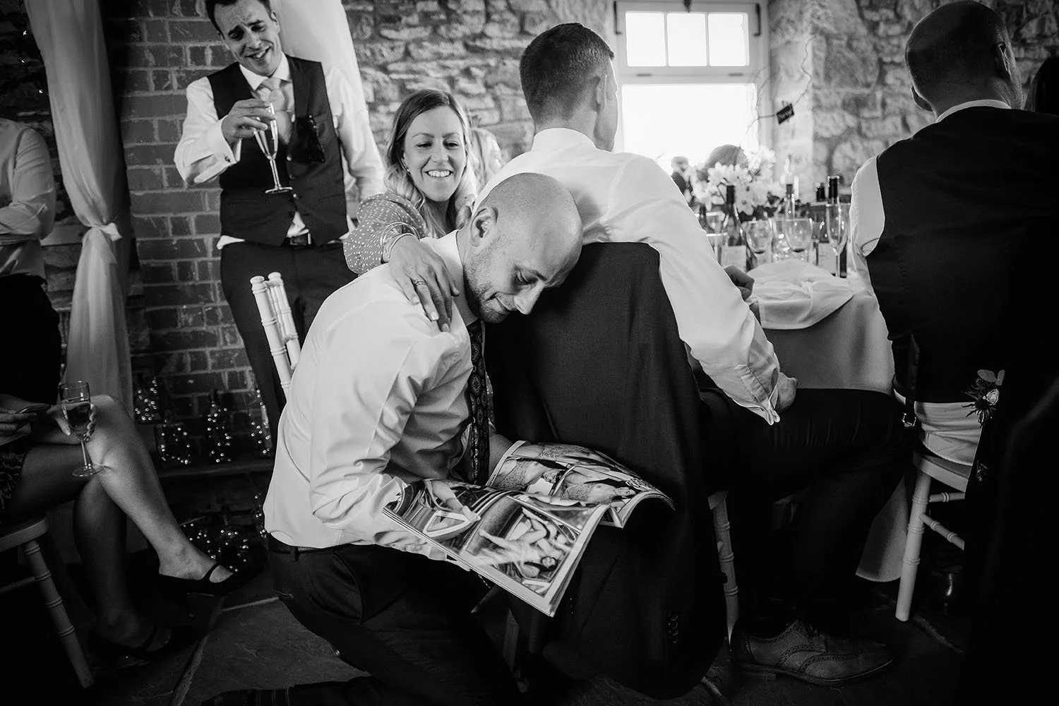Northside Farm wedding: groom resting his head on a guest's shoulder while another reads a magazine, black and white