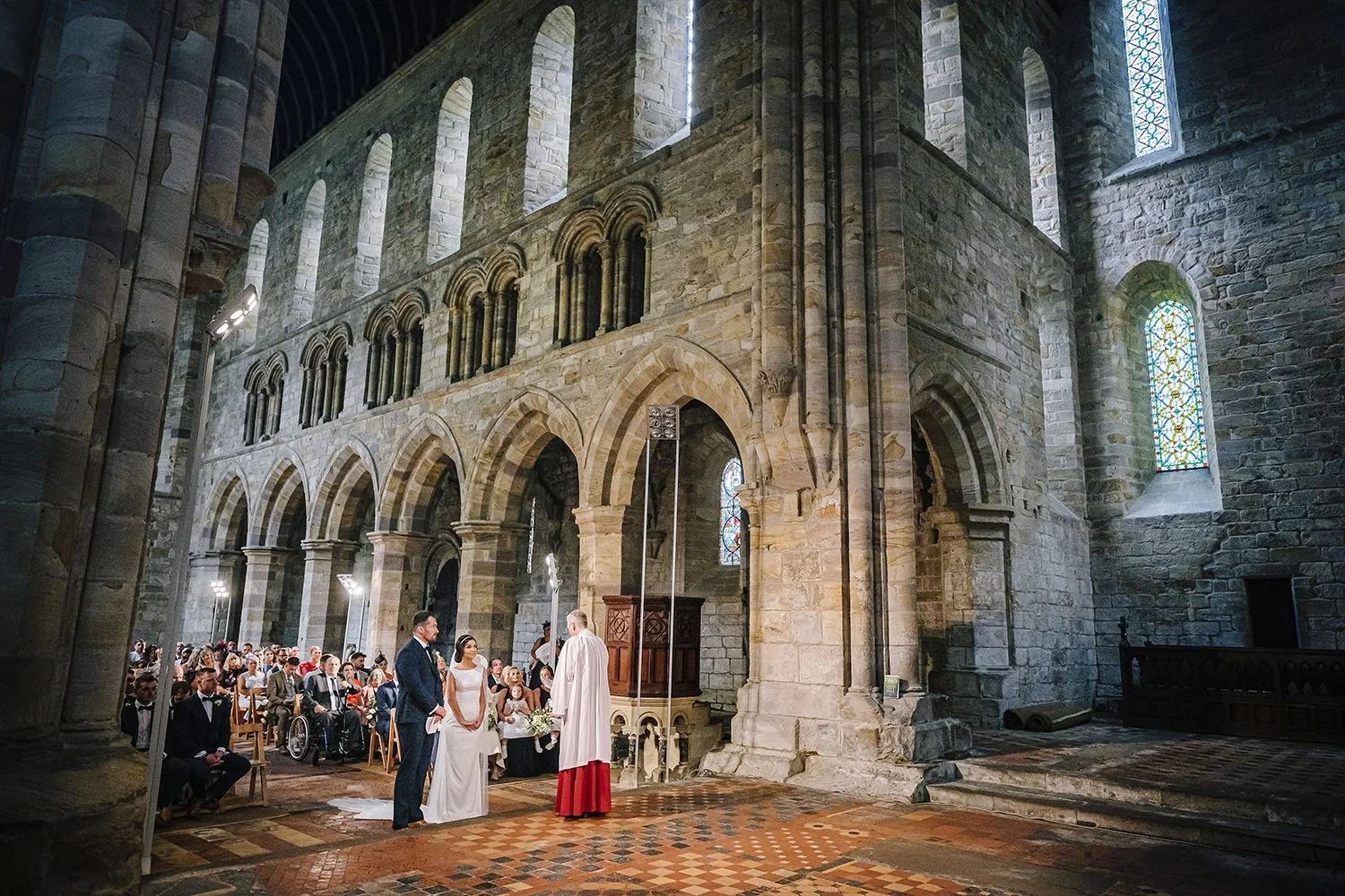 Brinkburn Priory wedding: wedding ceremony in a large stone abbey with tall arches, the vicar in red robes