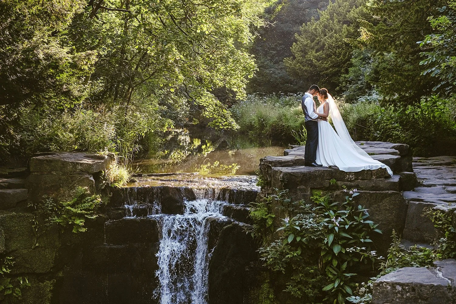 Jesmond Dene House wedding: bride and groom kissing on a stone ledge above a small waterfall in golden woodland light.