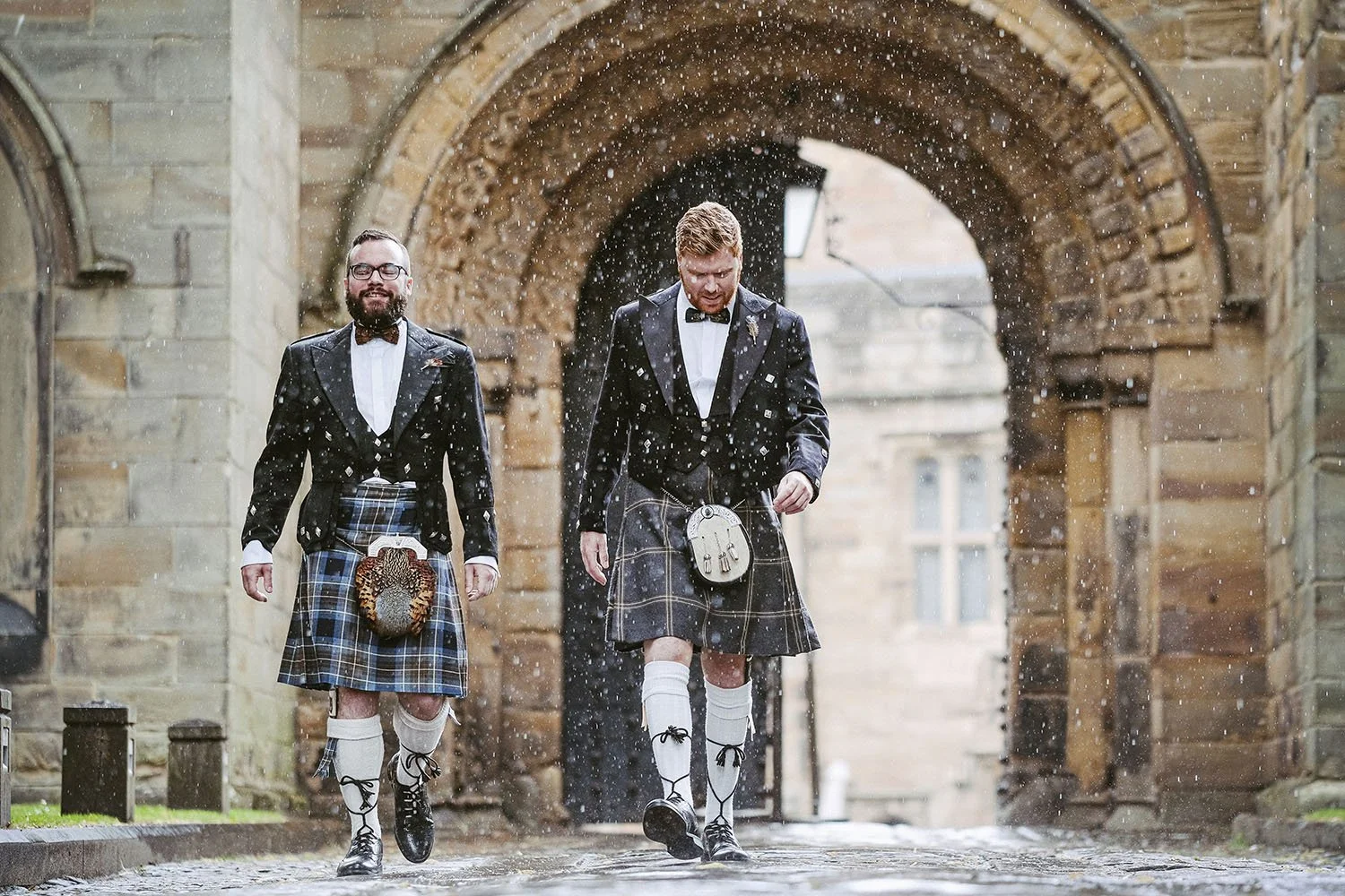 Durham Castle wedding: groom and best man in kilts walking through a stone archway as confetti falls.