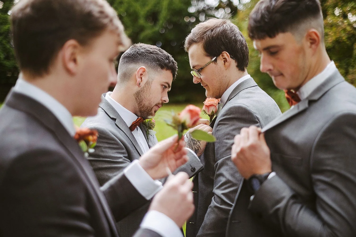 Groom and groomsmen pinning flowers onto each other's grey suits in a garden.