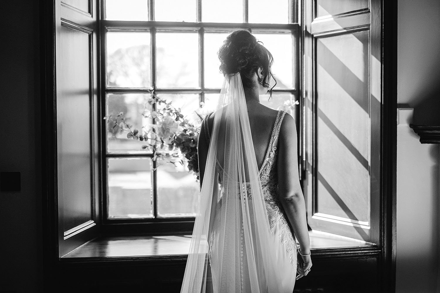 Black-and-white photo of a bride in a wedding dress and veil standing by a sunlit window