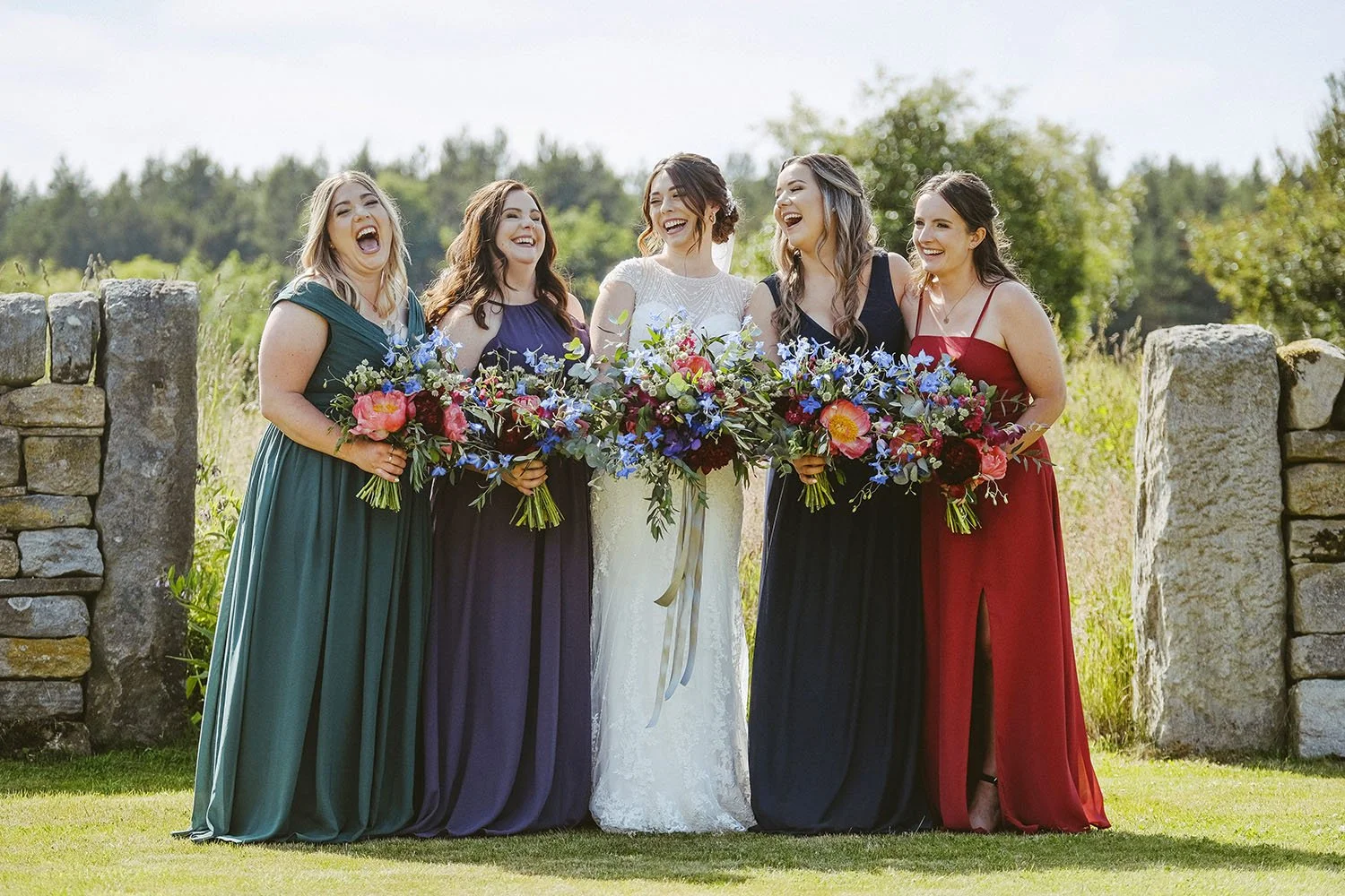 Healey Barn wedding: bride and bridesmaids laughing with colourful bouquets in front of a stone wall.