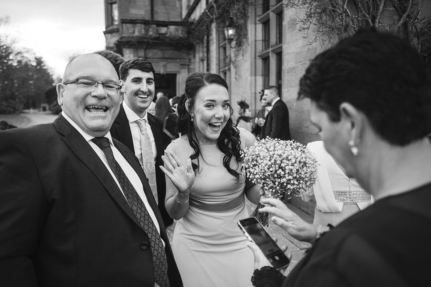 Ellingham Hall wedding: guests laughing and waving, a bridesmaid holding a gypsophila bouquet, in black and white.