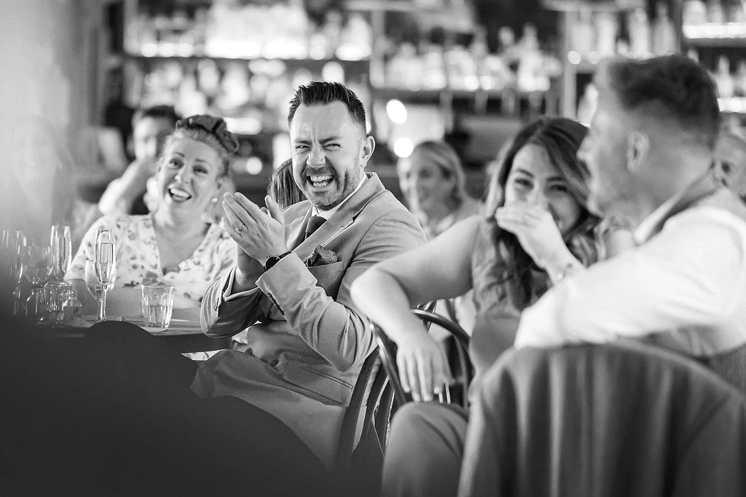 Healey Barn wedding: guests laughing and clapping at a table in front of a bar, in black and white.
