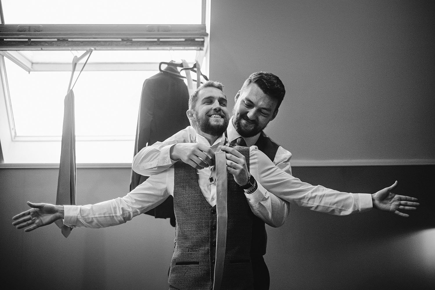 Ellingham Hall wedding: groomsman adjusting the groom's tie while the groom stretches his arms wide, in black and white.