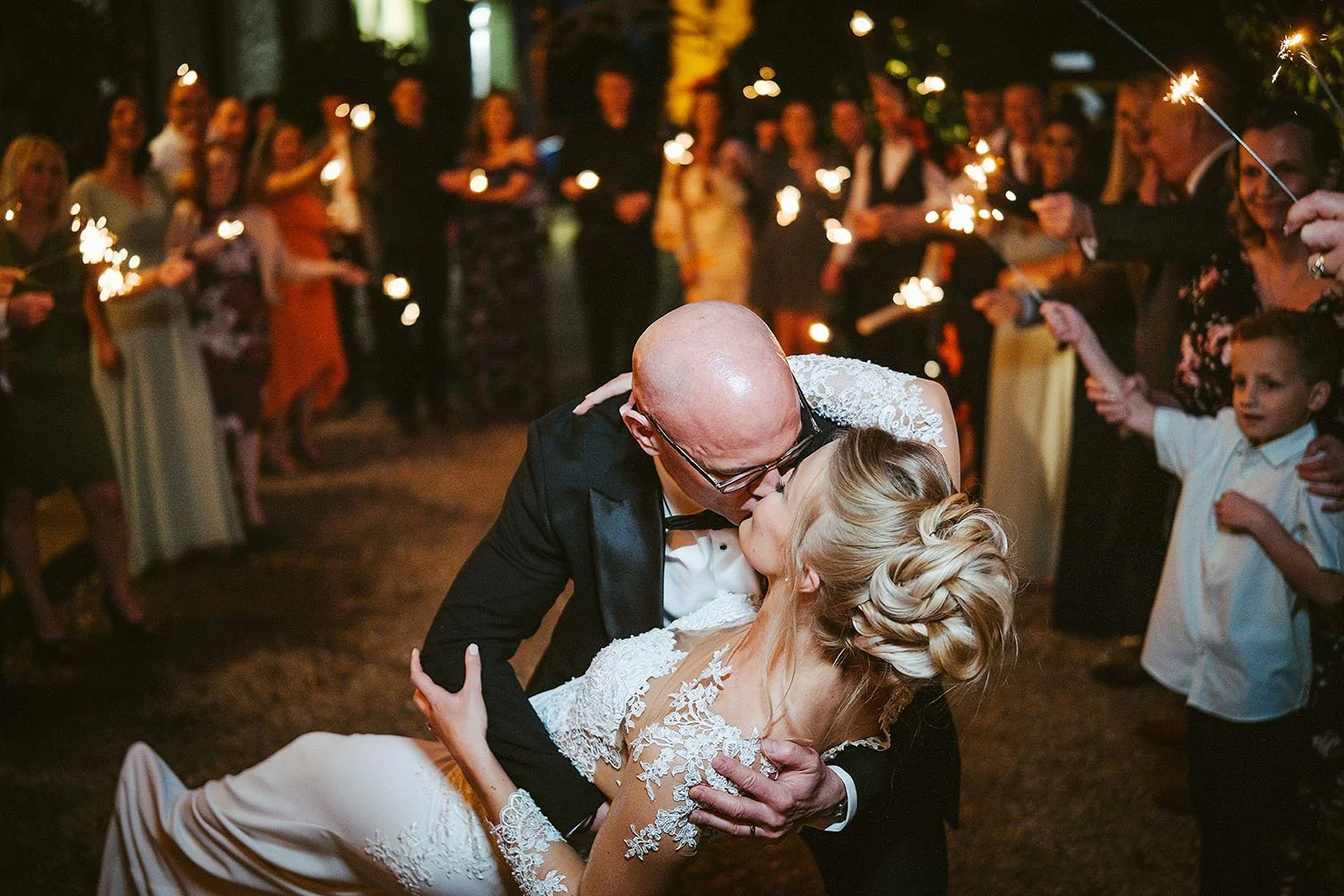 The Croft Hotel wedding: groom dipping the bride for a kiss at night as guests holding sparklers and a young boy look on.