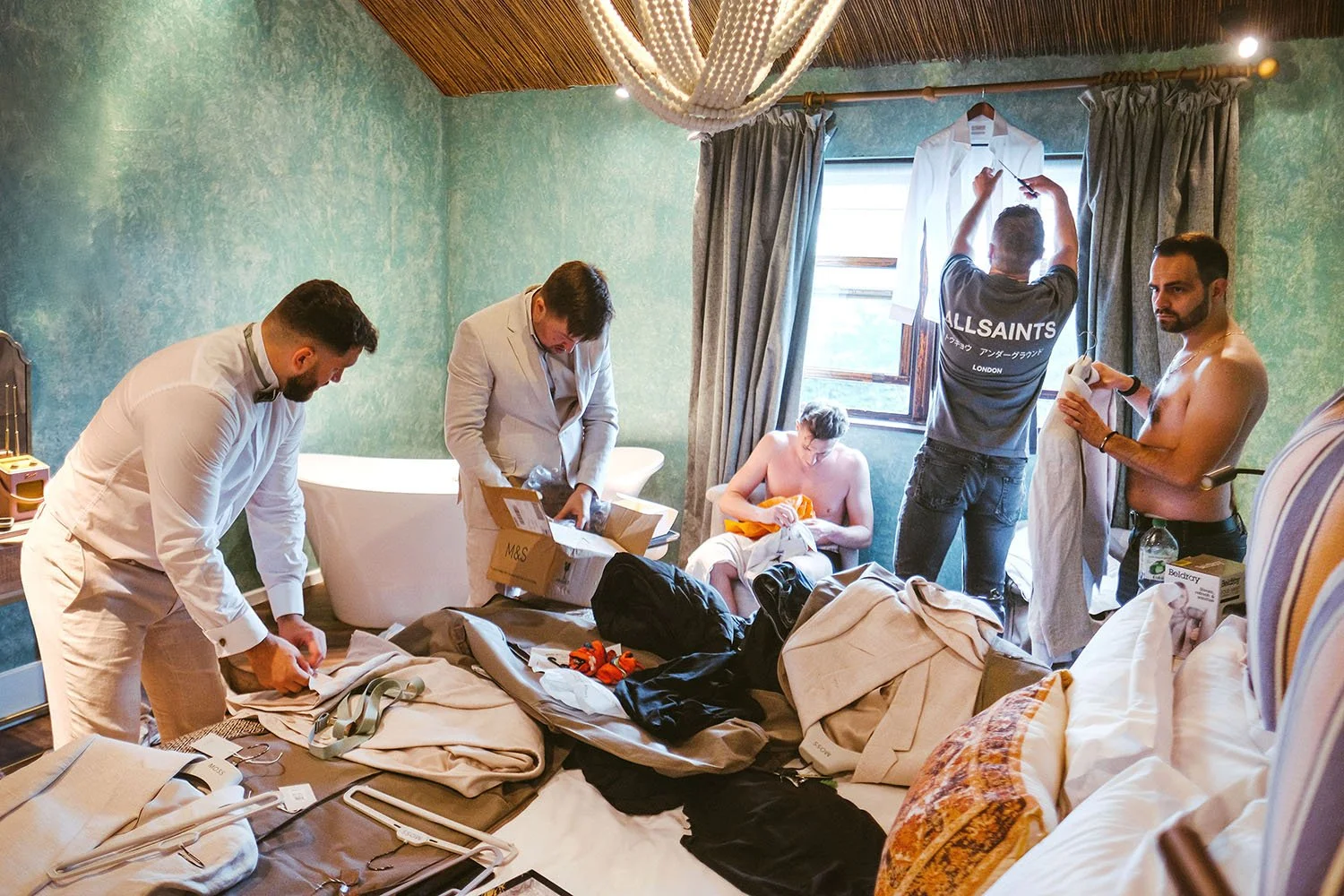 Runa Farm wedding: groom and groomsmen unpacking suits and clothes in a teal bedroom, some half-dressed and laughing.