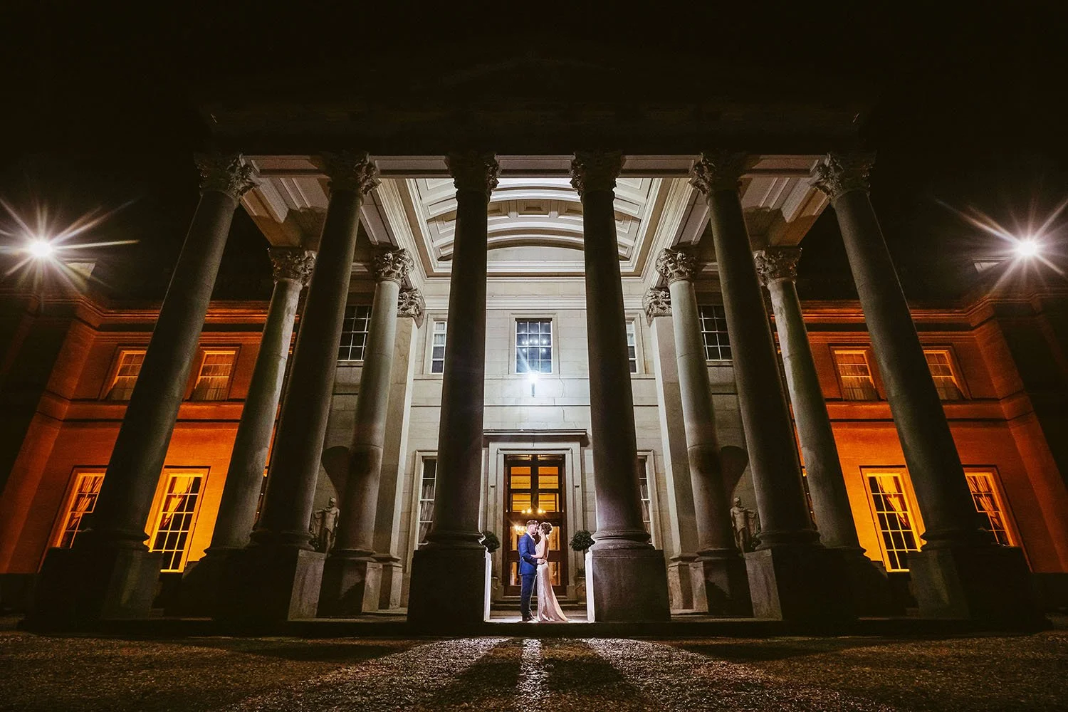Wynyard Hall wedding: bride and groom kissing in a lit doorway beneath tall stone columns at night