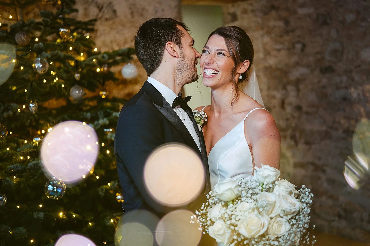 Bride and groom smiling beside a decorated Christmas tree, with the bride holding a white rose bouquet.