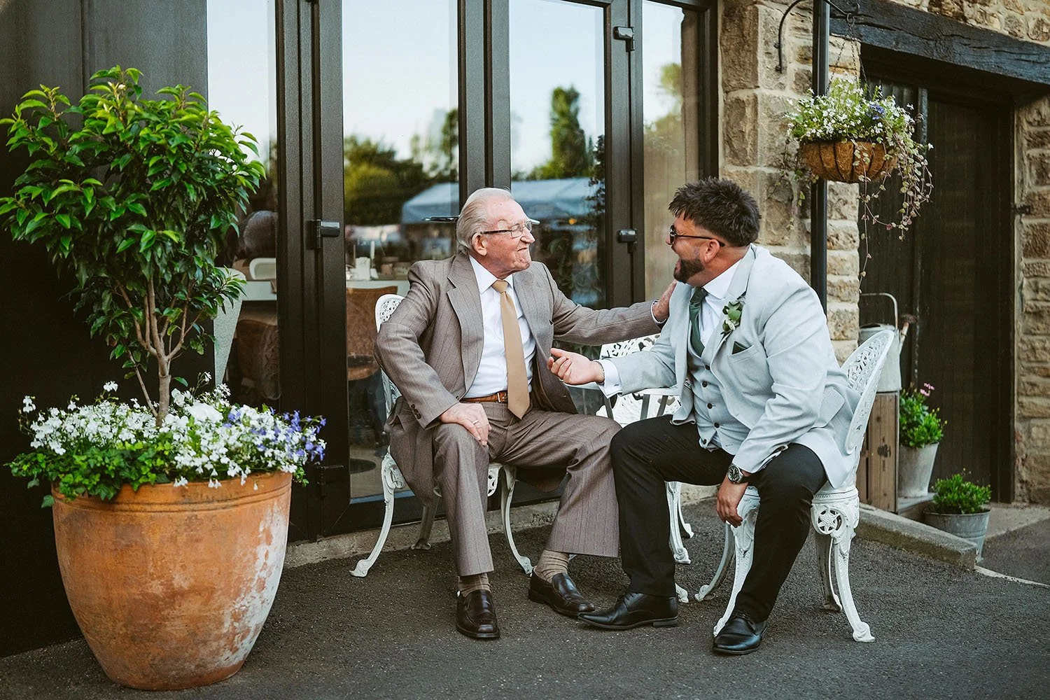 South Causey Inn wedding: groom in a pale blue suit and his father in a tan suit laughing together on white garden chairs