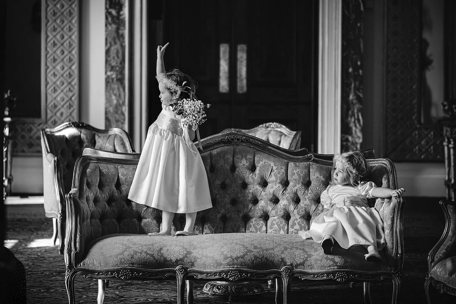 Wynyard Hall wedding: two flower girls playing on an antique sofa in a grand room, one raising her hand, black and white