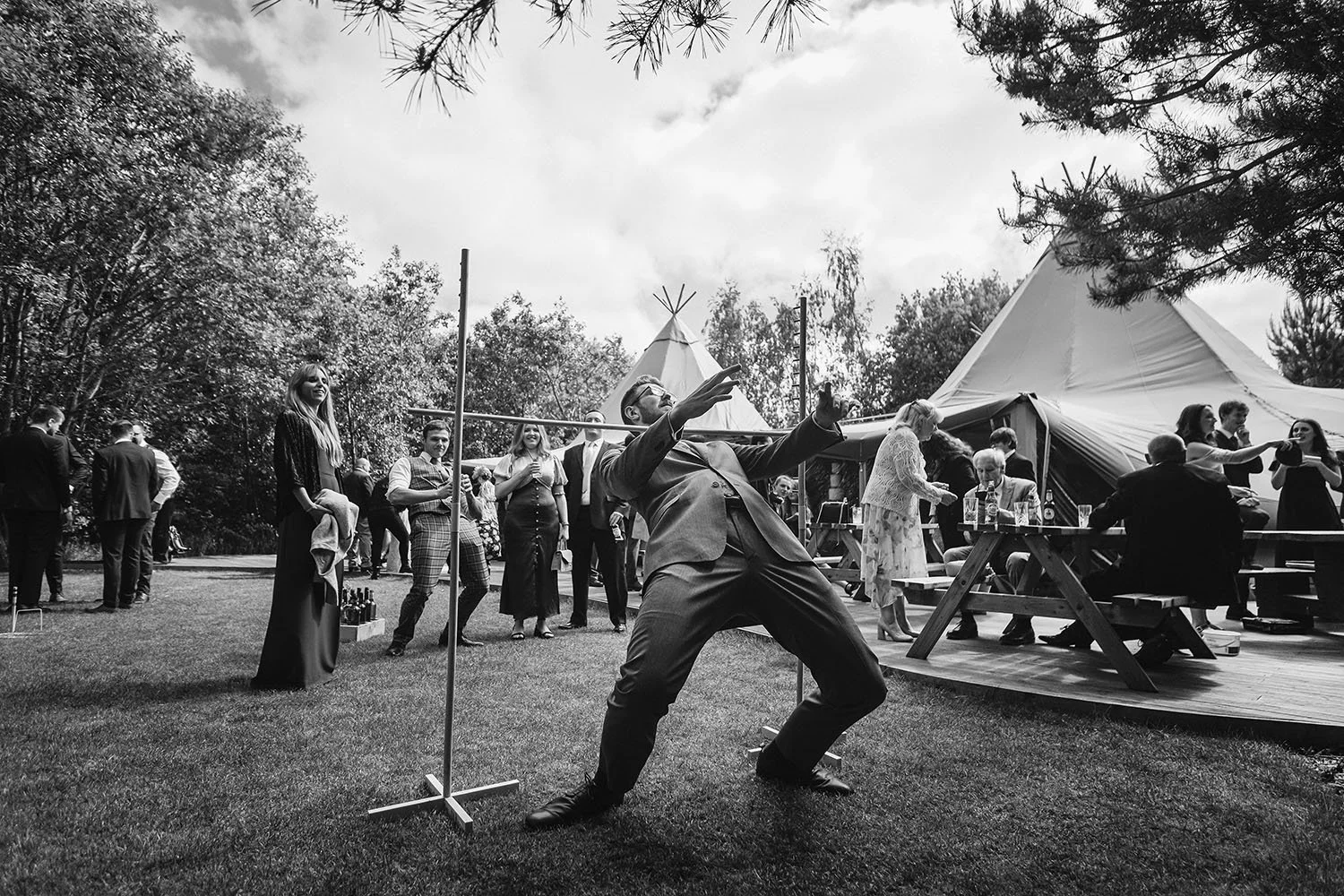 Bradbury Glade wedding: guest leaning back under a limbo bar on the lawn as others watch near a tipi, in black and white.