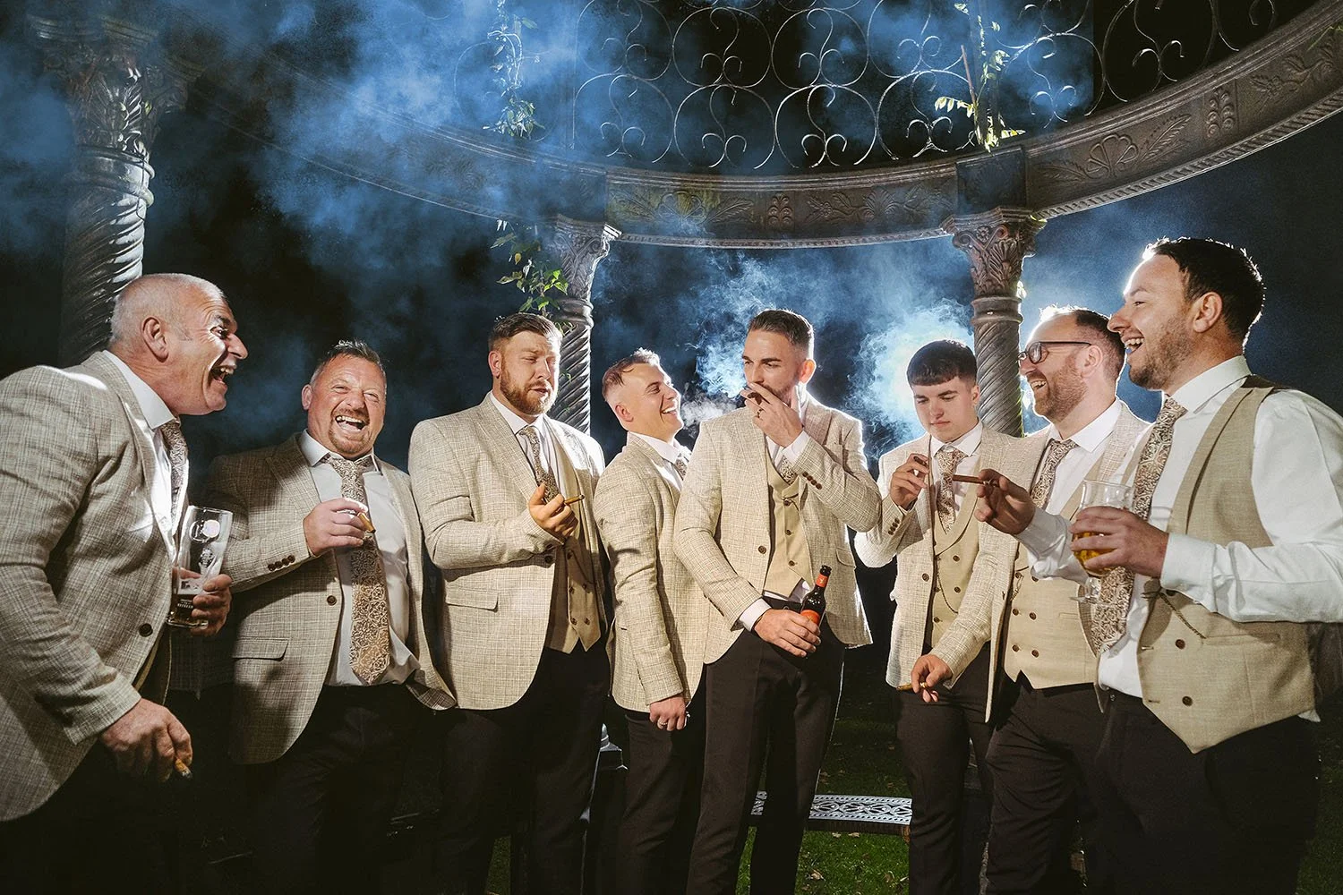 Beamish Hall wedding: groomsmen laughing and holding cigars under a stone gazebo at night.