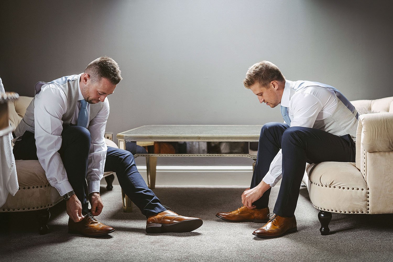 Ellingham Hall wedding: groom and best man sitting on cream sofas putting on brown brogues while getting ready.