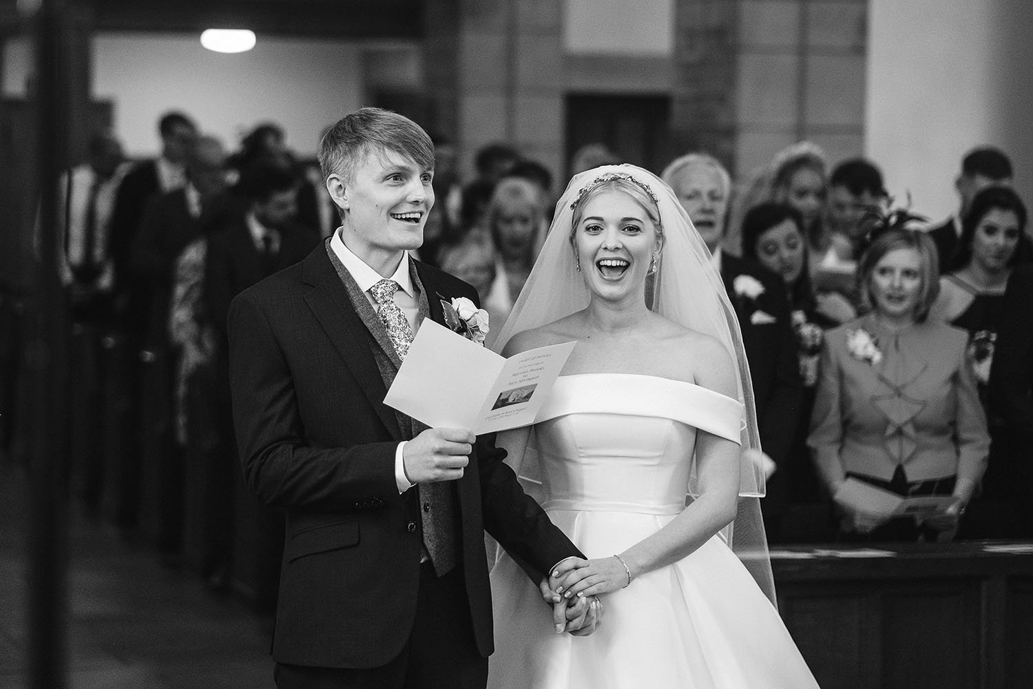 Durham School wedding: bride and groom laughing during the ceremony as the groom holds an order of service, in black and white.