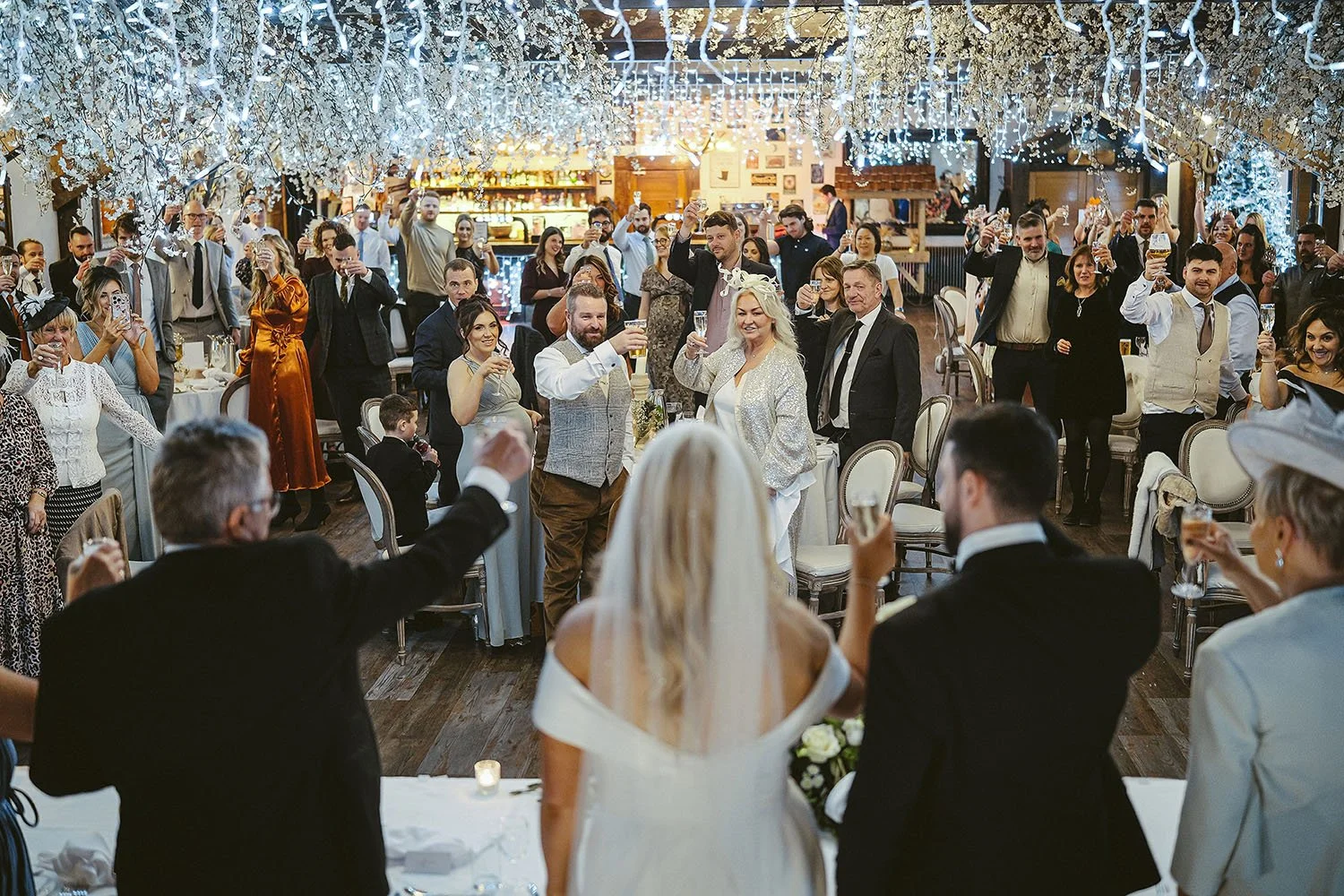 South Causey Inn wedding: guests cheering and raising glasses as the bride and groom enter a room hung with fairy lights.