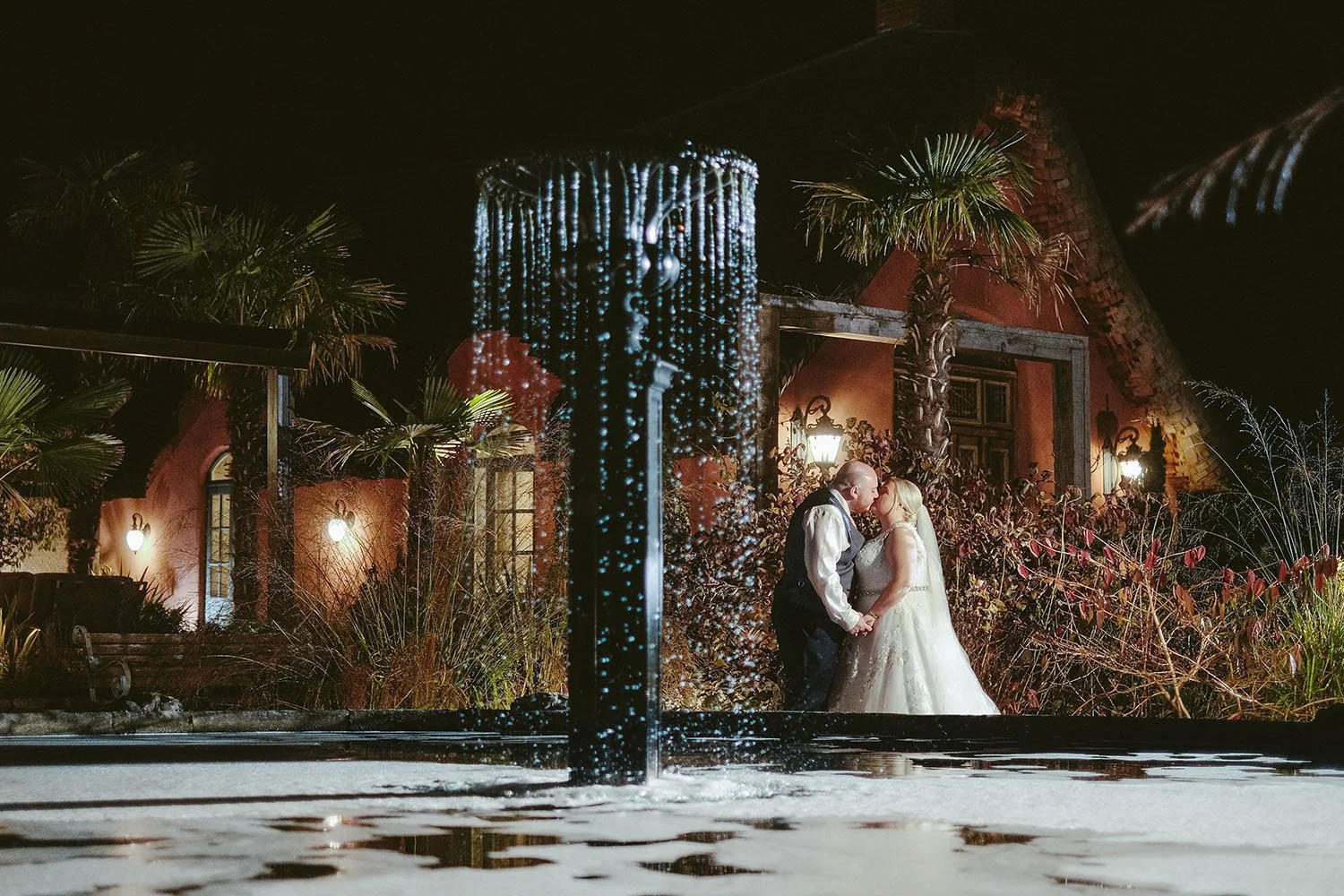 Le Petit Chateau wedding: bride and groom kissing at night behind a fountain in a lantern-lit garden.