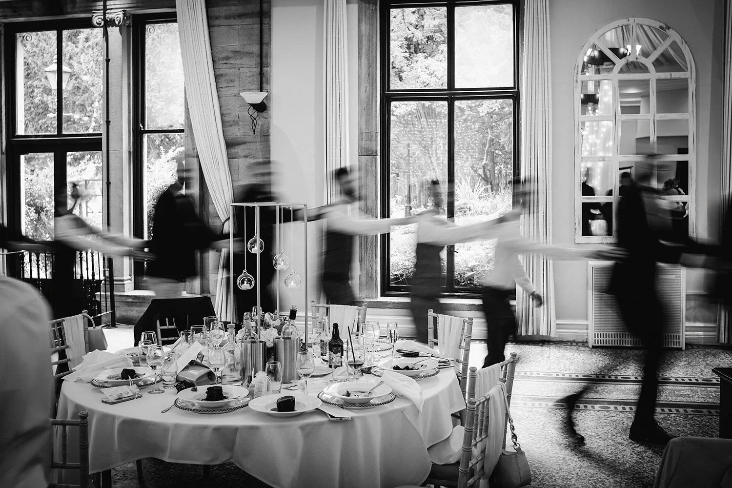 Black-and-white photo of a formal dining room with a set table and motion-blurred guests walking around it.