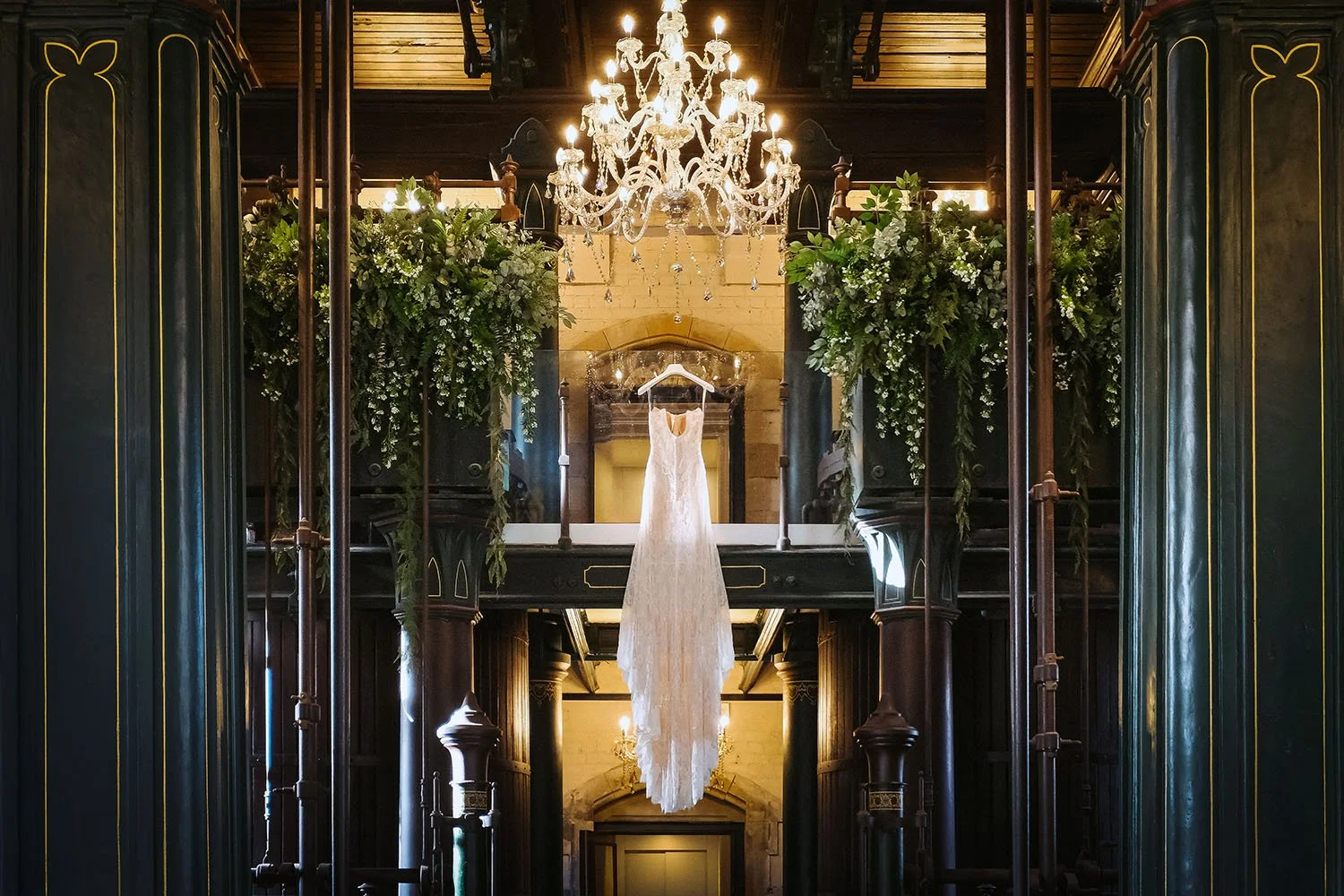 Dalton Pump House wedding: wedding dress hanging beneath a chandelier between dark green columns and hanging foliage.