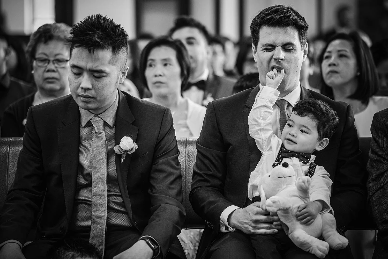 Black-and-white wedding ceremony photo of guests seated, including a yawning man holding a young boy with a stuffed animal