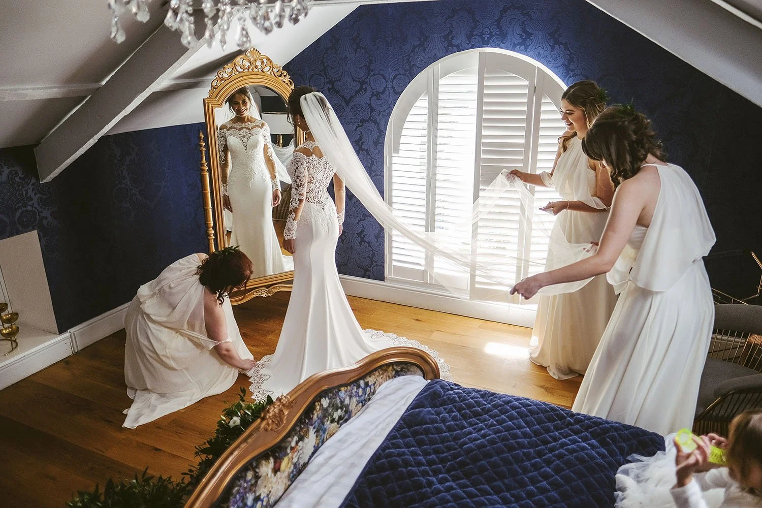 Woodhill Hall wedding: bride in front of a gold mirror in a blue attic bedroom as bridesmaids arrange her veil and train