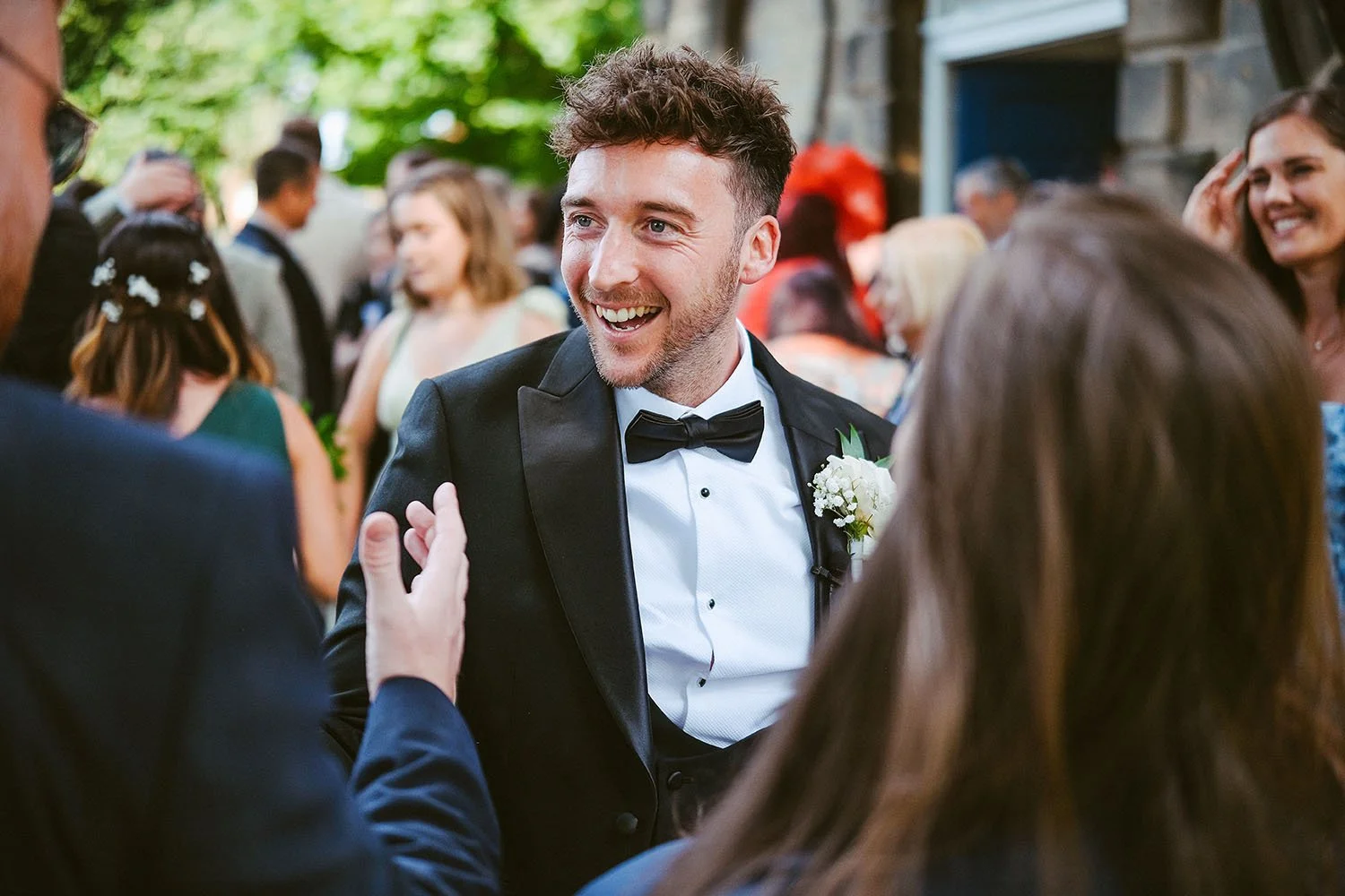 Groom in a tuxedo and bow tie laughing as guests crowd round him in a garden.