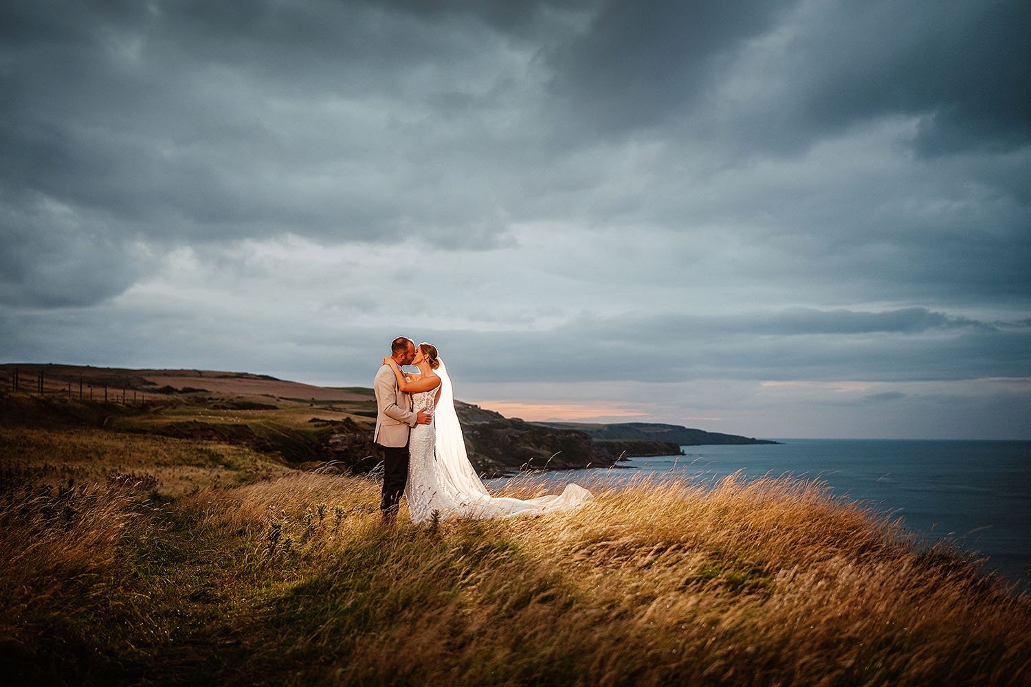 Marshall Meadows Manor House wedding: bride and groom kissing on a grassy clifftop above the sea under a dramatic cloudy sky.
