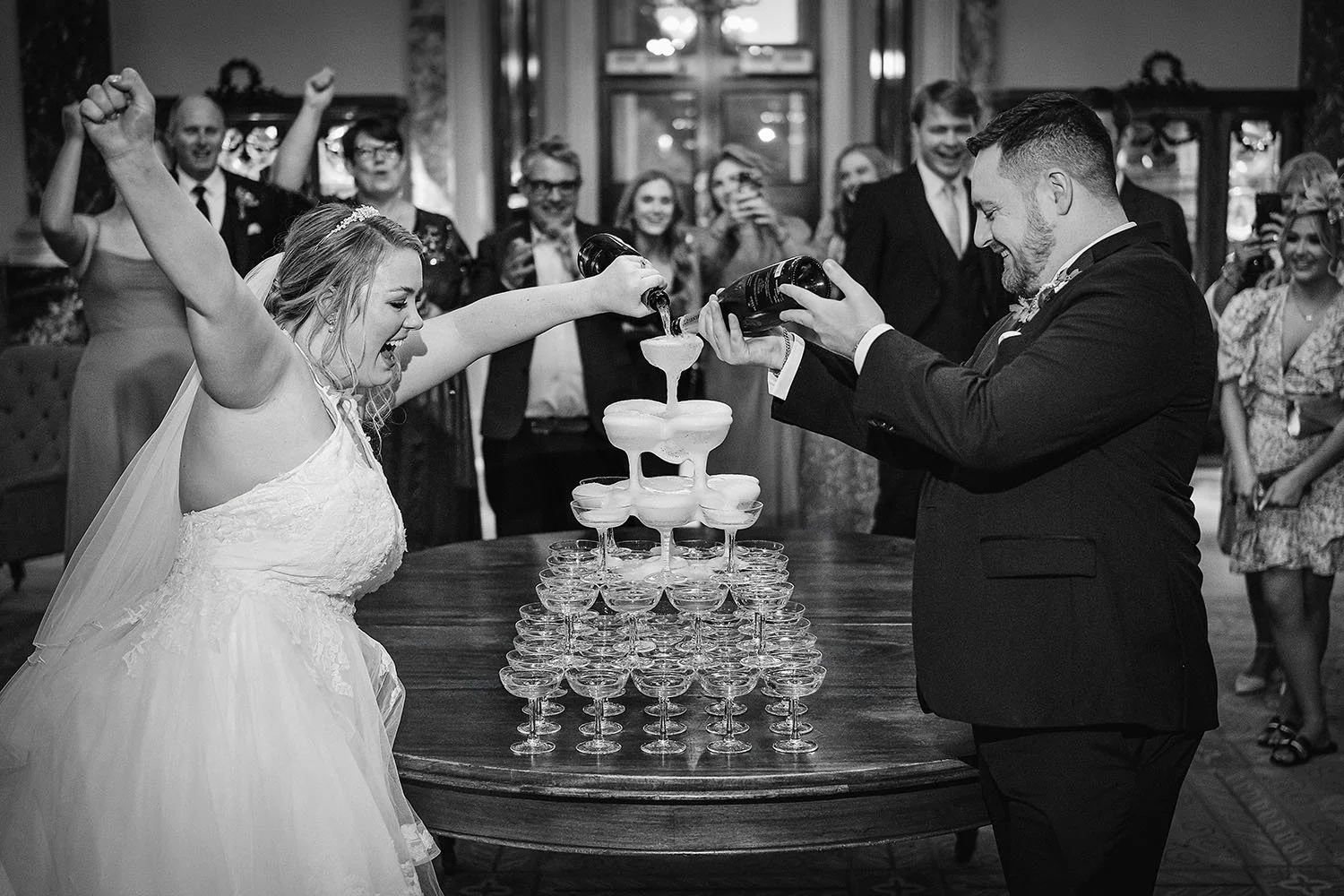 Wynyard Hall wedding: bride cheering with her arm raised as the groom pours champagne over a coupe glass tower