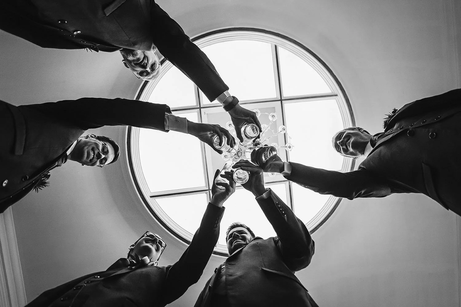 Black-and-white low-angle photo of five men in suits toasting with drinks beneath a circular skylight
