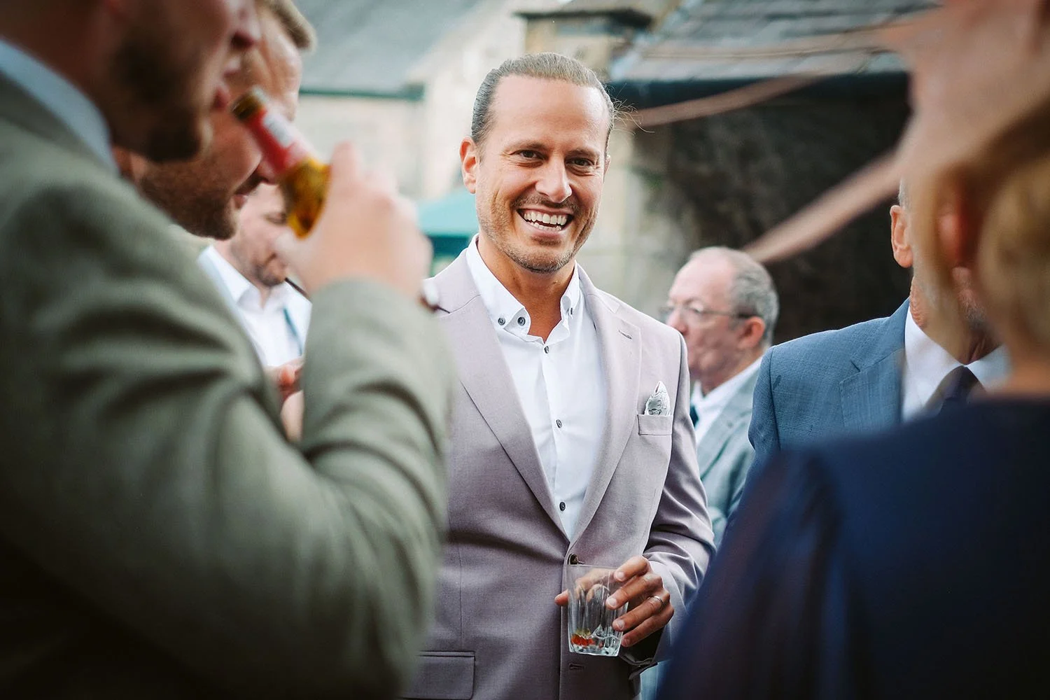 Healey Barn wedding: smiling man in a lilac jacket with a tied-back ponytail holding a drink among guests at a garden reception.