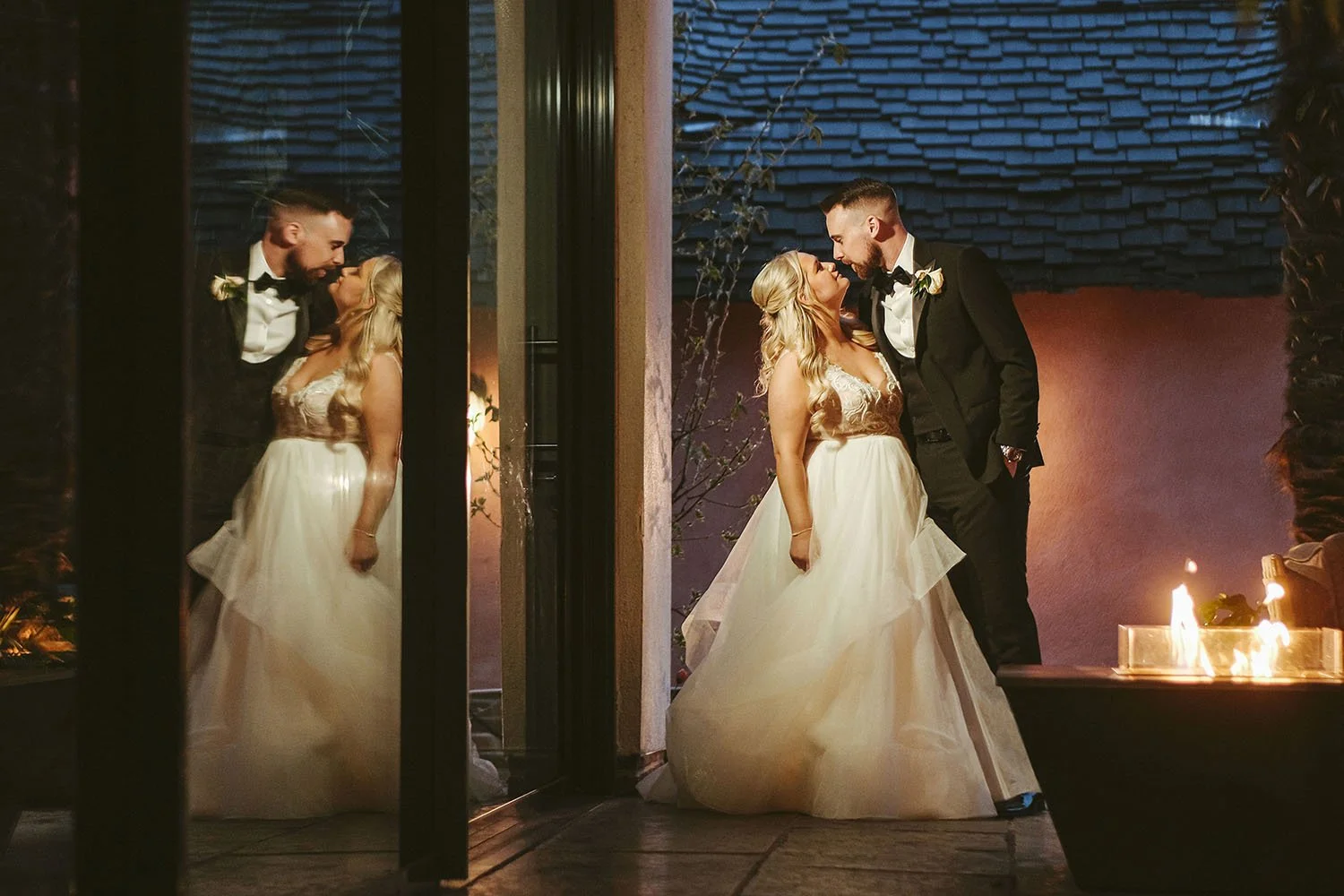 Le Petit Chateau wedding: bride and groom kissing by a fire pit, seen through glass doors with a mirrored reflection.