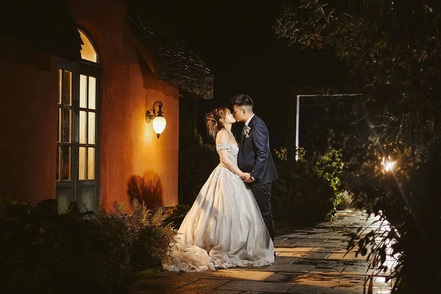 Le Petit Chateau wedding: bride and groom kissing on a stone path at night beside a warmly lit orange wall.