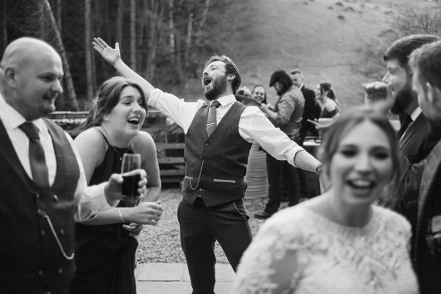 Bradbury Glade wedding: man with arms outstretched singing as wedding guests laugh around him, in black and white.