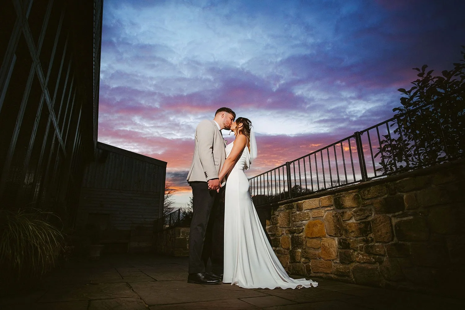 South Causey Inn wedding: bride and groom kissing on a stone terrace under a vivid purple and pink sunset sky.
