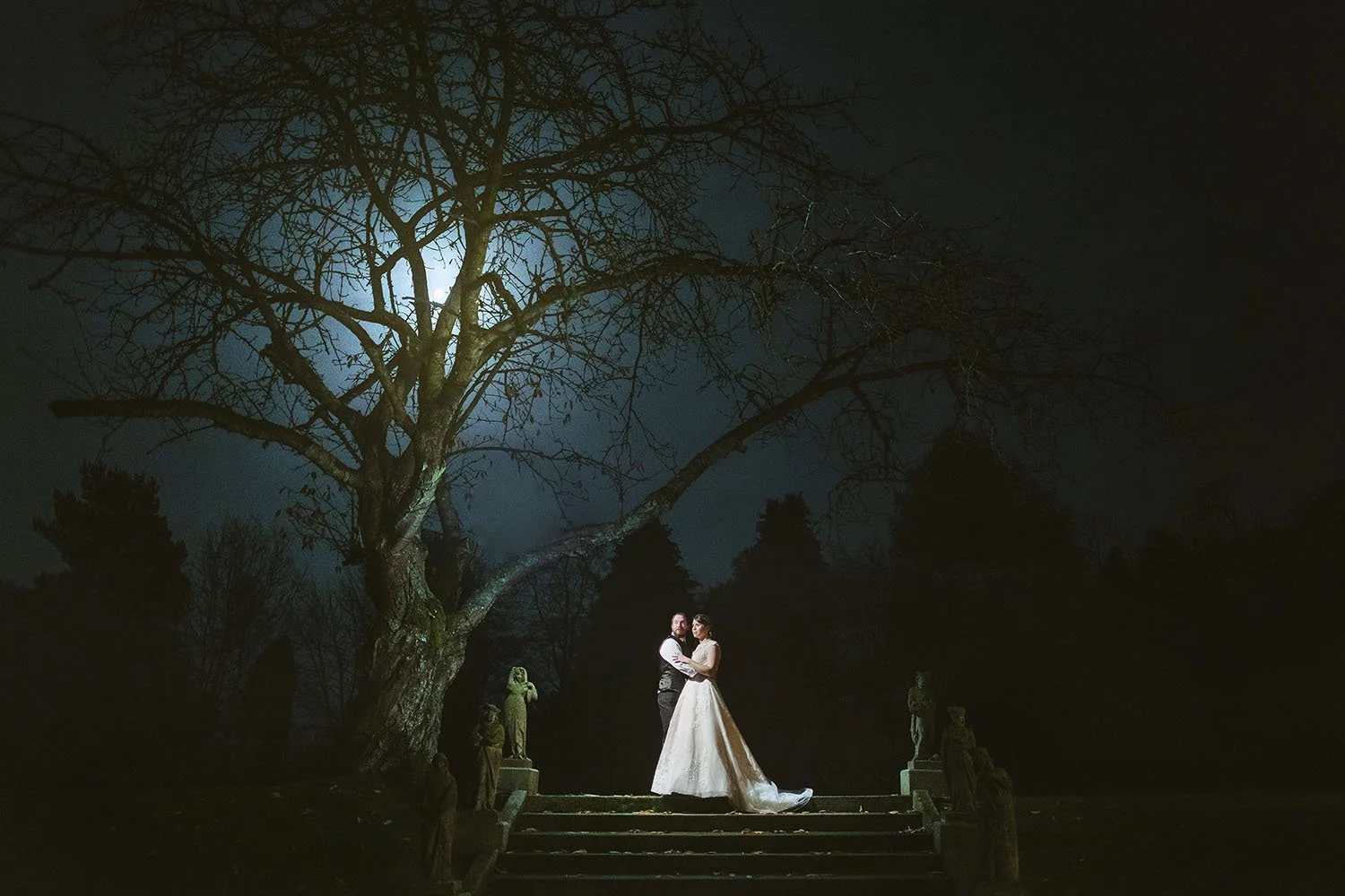 Lartington Hall wedding: bride and groom embracing on stone steps beneath a large moonlit tree at night